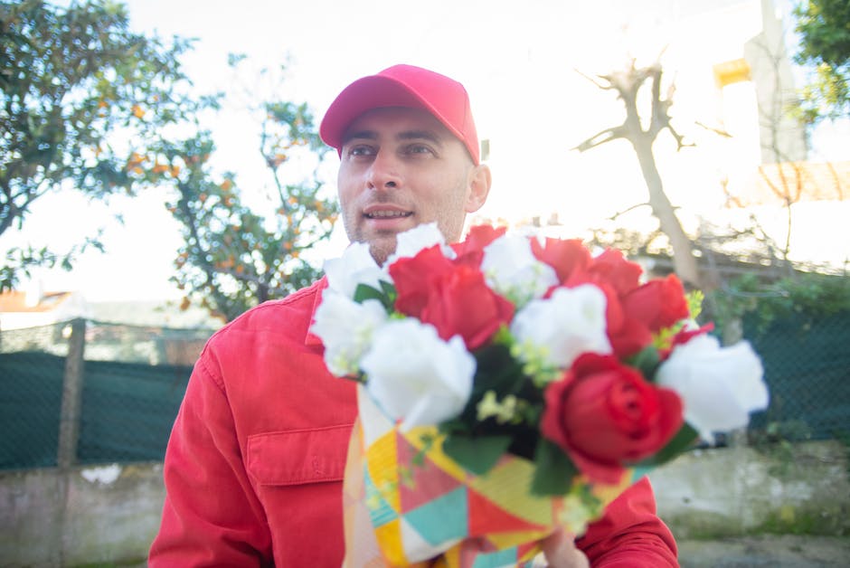 A man dressed in a red cap and matching red coat is holding a vibrant bouquet of fresh flowers, including red and white roses, wrapped in colorful paper. He is outdoors, with a background of trees, a 