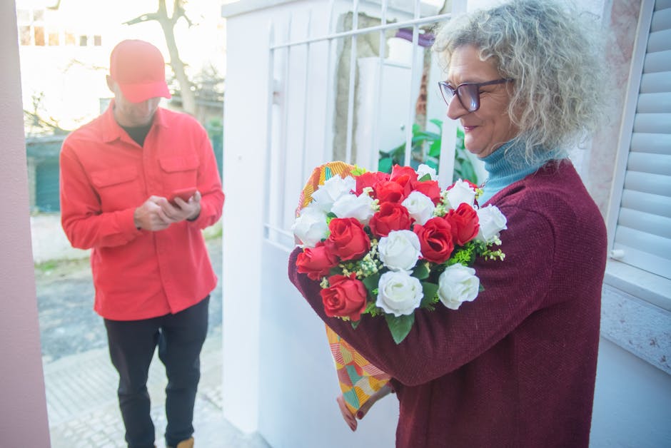 A woman with curly gray hair, glasses, and a maroon sweater holds a vibrant bouquet of fresh red and white roses wrapped in patterned paper near her chest, smiling softly as she prepares for a flower 