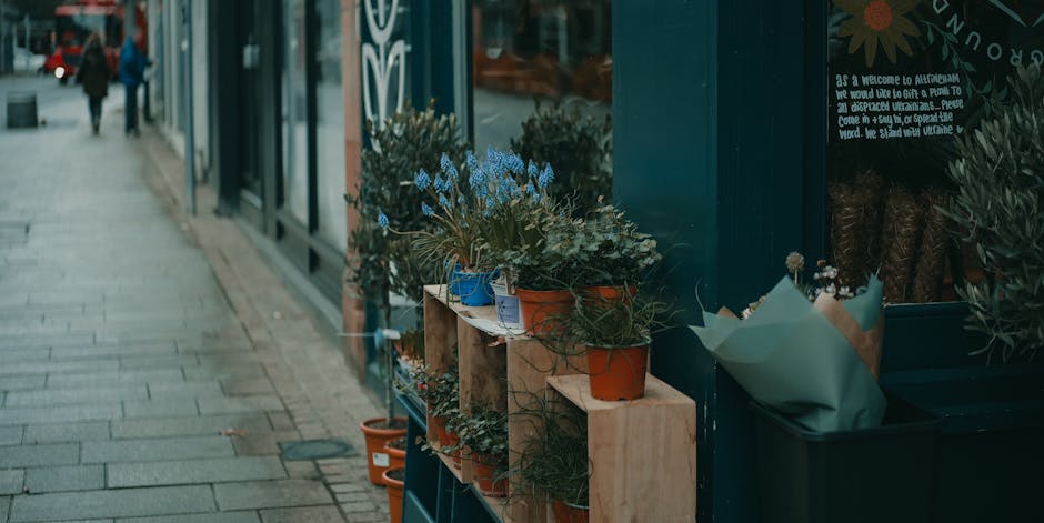 A display of potted plants and flowers arranged on wooden shelves outside a flower shop, including blue, orange, and terracotta pots with greenery and small flowering plants. The arrangement is set ag