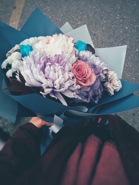 A hand holding a bouquet of fresh flowers wrapped in dark and light blue paper, featuring a large white chrysanthemum, soft pink rose, purple flowers, and smaller white blooms with green stems. The bo