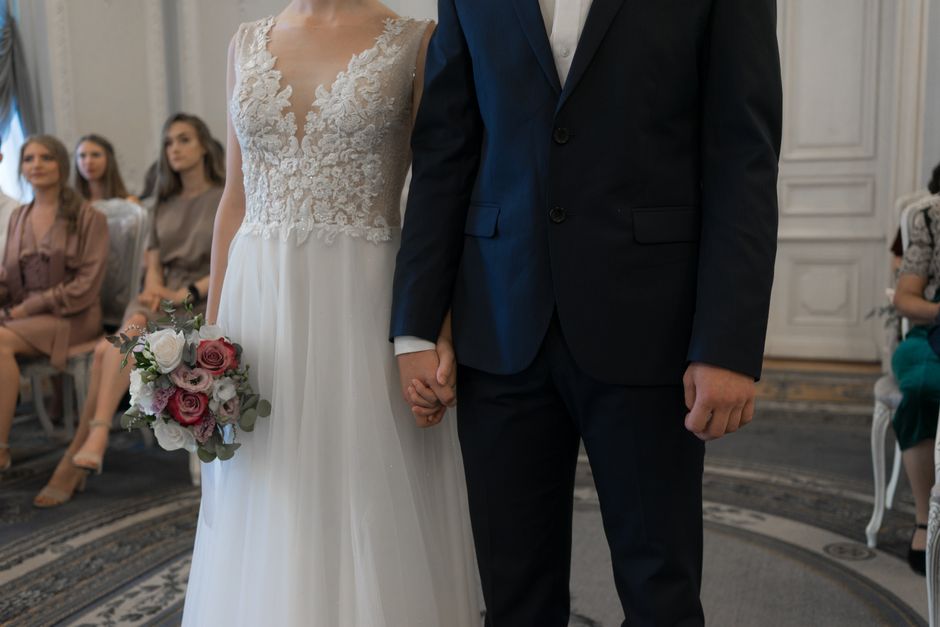 A bride in a lace bodice wedding dress holding a bouquet featuring pink, white, and blush roses with green foliage, standing next to a groom in a dark blue suit with a white shirt, holding hands durin
